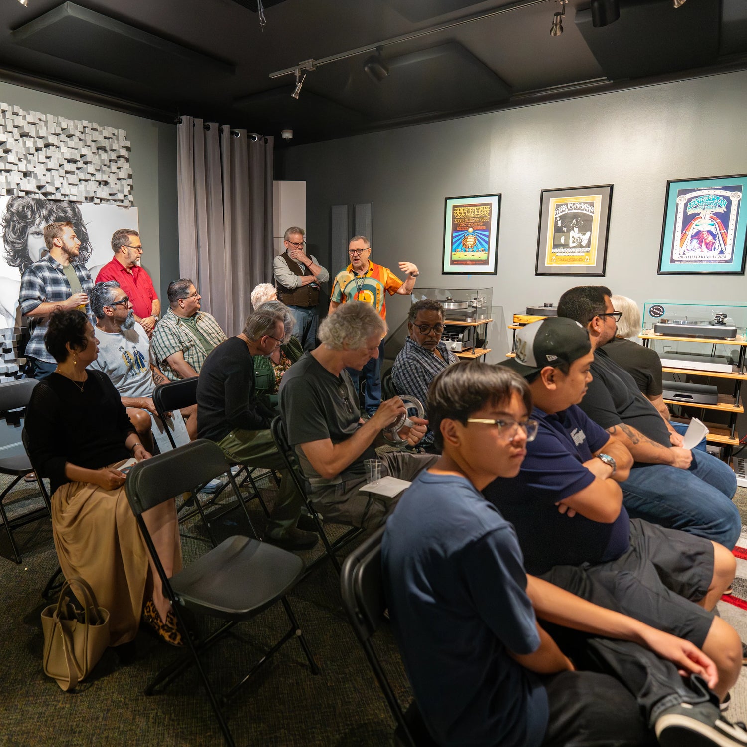 A group of guests sit in folding chairs inside the Upscale Audio listening room while a presenter stands at the front giving a talk. Turntables and framed posters line the wall, and acoustic treatment panels are visible near the ceiling and back corner.